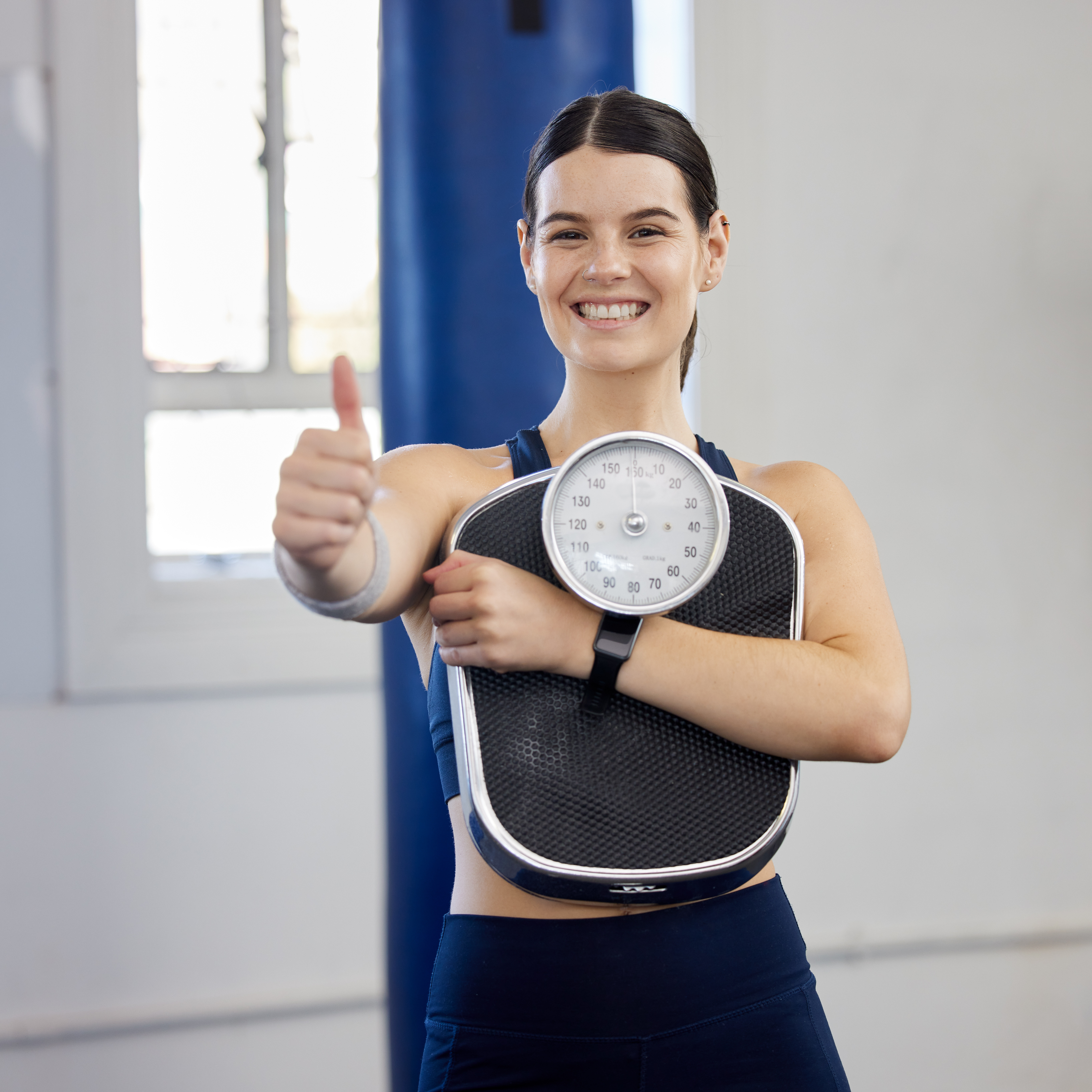 Woman, gym portrait and thumbs up with scale, wellness and weight loss goal for health, body or fit